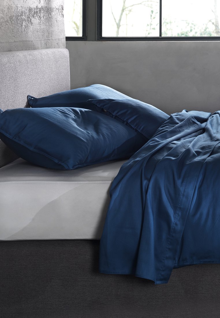 Postel s šedým čelem, tmavě modrými polštáři a ladící tmavě modrou přikrývkou, která zčásti zakrývá bílou matraci poblíž okna.