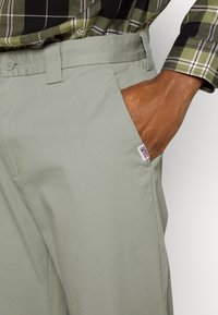 Light green, cotton pants with a pocket detail, a woven label on the side seam, and a smooth texture. Paired with a black and green plaid shirt.