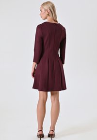 Bordeauxfarbenes A-Linien-Kleid mit Dreiviertelärmeln, Reißverschluss im Rücken und dezenten Faltendetails. Glatte Textur und tailliertes Oberteil, das in einen ausgestellten Rock übergeht.