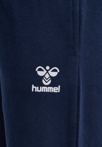 Granatowy materiał o miękkiej teksturze, z białym logo "hummel" z stylizowanym wzorem pszczoły i pionowym przeszyciem.