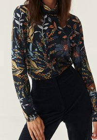 Kvinna som bär en marinblå polotröja med flerfärgade blommönster instoppad i högmidjade mörka byxor, står med händerna nära höfterna.