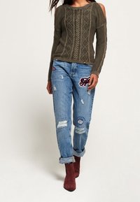 Olivově zelený svetr s odkrytými rameny a strukturovaným pleteným vzorem, kombinovaný s popraskanými modrými džíny s ná�šivkami a ohrnutými nohavicemi.