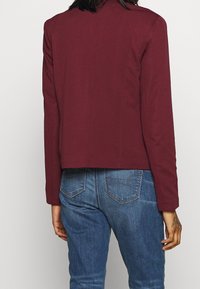 Personne portant une veste à manches longues bourgogne et un jean en denim bleu, vue de dos avec un tatouage visible sur la main droite.