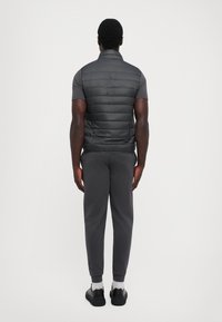 Gilet matelassé gris foncé, sans manches, avec une texture lisse. Assorti à un pantalon long et des baskets noires. Design minimaliste.