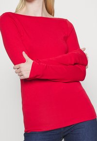 Maglia rossa a maniche lunghe con scollo a barca, realizzata in un materiale morbido e elasticizzato. Design aderente con polsini allungati. Indossata con jeans di denim.