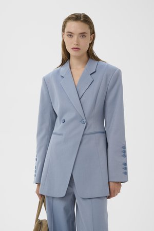 Kvinde med lige blondt hår iført oversize lyseblå blazer og matchende bukser, holder en tanfarvet håndtaske mod en ensfarvet baggrund.