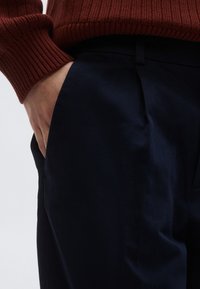 Παντελόνια από ύφασμα ναυτικού μπλε με επίπεδη μπροστινή όψη και πλαϊνές τσέπες, σε συνδυασμό με ένα πουλόβερ σε σκουριά απόχρωση με ανάγλυφες ρίγες. Η υφή φαίνεται λεία και καθημερινή.