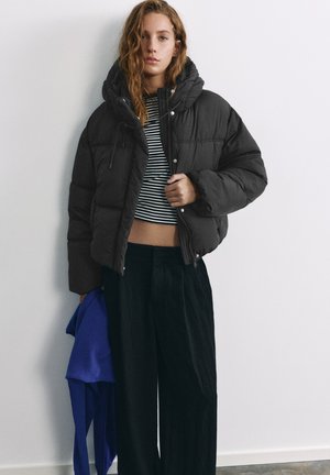 Un mannequin porte une doudoune noire sur un haut court rayé et un pantalon à jambes larges, et tient un vêtement bleu devant un mur uni de studio.