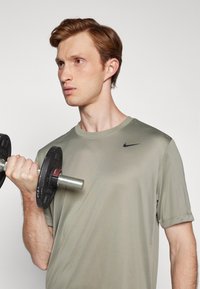 Tricou sportiv cu mâneci scurte, de culoare verde măslin, cu un logo Nike negru, ținut de o persoană care ridică o bară. Textură netedă, design ajustat.