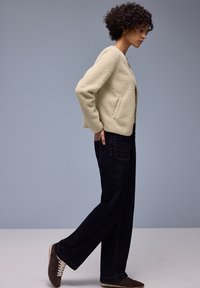 Vrouw in een beige fleecejack, donkere wijde spijkerbroek en bruine sneakers loopt zijwaarts tegen een effen grijze achtergrond.