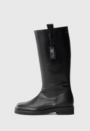 Bota de cuero negro hasta la rodilla con punta redondeada, suela plana y un tirador. Presenta una etiqueta con detalles numéricos adjunta. Textura suave.