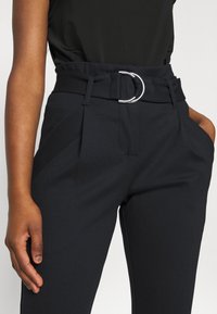 Pantalons noirs taille haute avec une large ceinture arborant une boucle circulaire en argent, en tissu texturé et avec des poches latérales. Coupe ajustée avec des plis à l'avant.