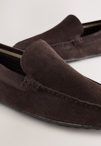Mocassins en daim marron avec une tige lisse et texturée, des détails cousus et une semelle en caoutchouc plate, conçus pour un usage décontracté.
