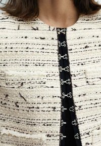 Gros plan sur une personne portant une veste en tweed crème et noire texturée avec des fermetures à crochet et œillets argentés le long de l'ouverture avant.