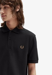 Svart polotröja gjord av strukturerad tyg, med en placket med två knappar och en liten tan laurel-logotyp på vänster bröst.