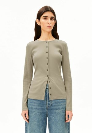 Jeune femme aux longs cheveux foncés portant un haut taupe à manches longues boutonné et un jean bleu ample, debout devant un fond blanc.