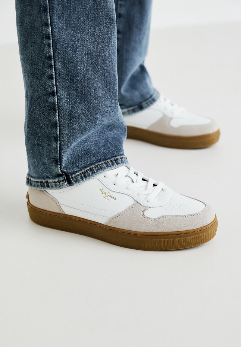 Baskets blanches et beiges avec semelles en gomme portées avec un jean bleu sur un fond clair.