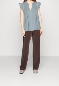 Top azzurro chiaro a maniche corte con dettagli a volant e scollatura a V, abbinato a pantaloni brown a gamba larga e ballerine grigie. Texture morbide in tutto.