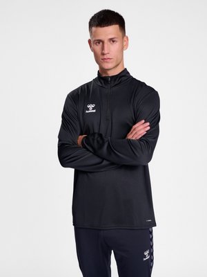 Jaunas vyras trumpais tamsiais plaukais, dėvintis juodą hummel sportinį megztinį ir kelnes, stovi sukryžiavęs rankas ant šviesaus vientiso fono.