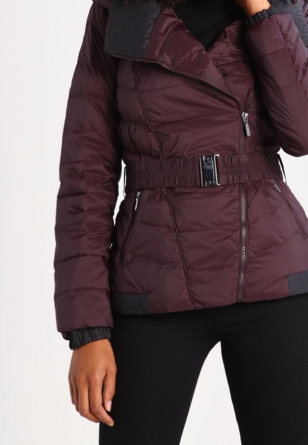 Femme portant une veste matelassée violet foncé avec une ceinture et une fermeture éclair, mains détendues, associée à un pantalon noir sur fond blanc.