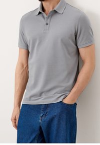 Polo shirt gris à manches courtes avec un col structuré, patte de boutonnage à trois boutons et un tissu texturé. Porté avec un pantalon en denim bleu.