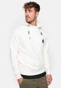 Sudadera blanca de tela texturizada, presenta un cuello alto, cuerdas ajustables y detalles de cuero. Llevada sobre una camiseta negra.