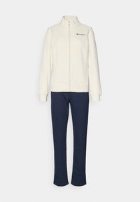 Hvit zip-up jakke med høy krage og svart logo, kombinert med marineblå joggebukser; begge laget av myk, teksturert stoff.