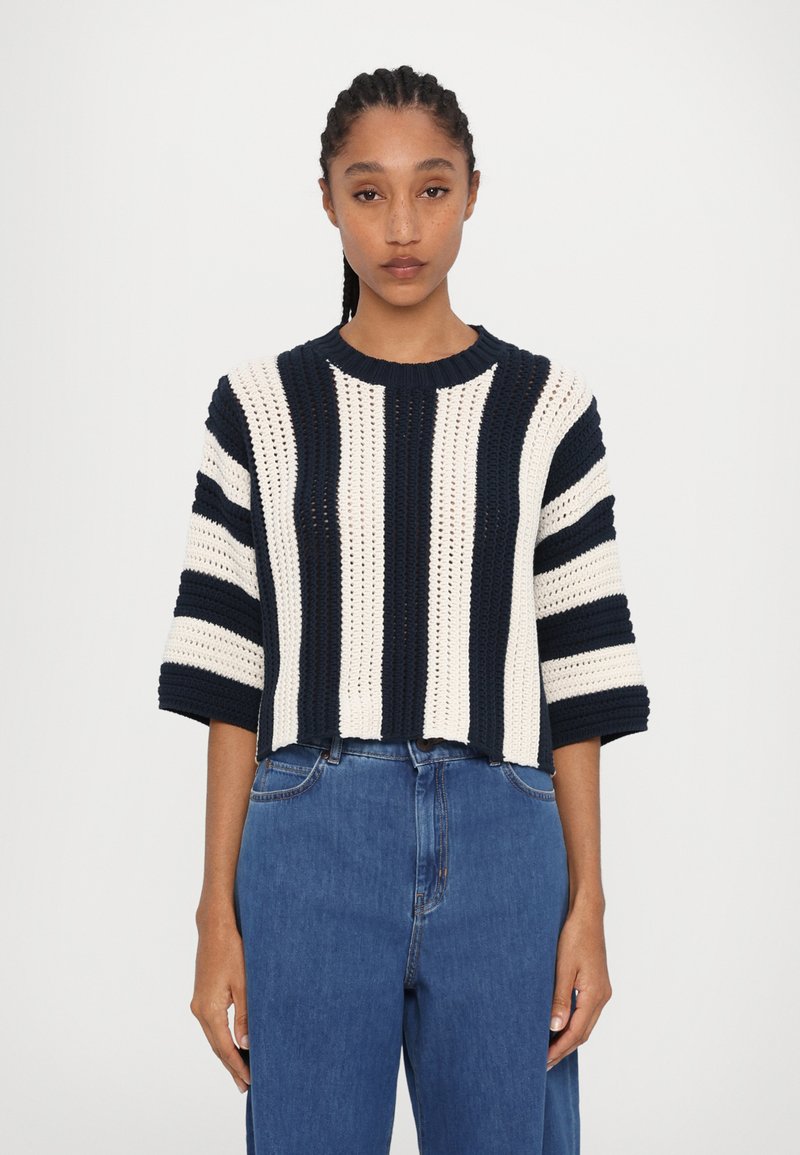 Pulover din tricot croșetat cu dungi orizontale bleumarin și crem, guler rotund, umeri căzuți și un design scurt. Asortat cu blugi albaștri.
