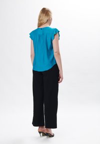 Türkise Kurzarmbluse mit Rundhalsausschnitt, gefaltetem Rücken und schwarzem Knopfdetail. Kombiniert mit schwarzen weiten Hosen und High Heels.