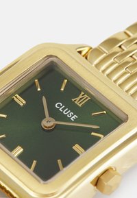 Montre carrée dorée avec un cadran vert, présentant des index horaires dorés, une couronne dorée et un bracelet en maillons métalliques. Le nom de la marque "CLUSE" est affiché.