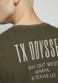 T-shirt vert olive avec un texte blanc imprimé au dos. Le texte comprend "TX ODYSSEY", "WAY OUT WEST" et des détails de localisation.