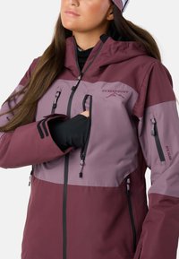 Giacca impermeabile bordeaux e viola con tasca sul petto con zip e tasche laterali, dotata di cappuccio e polsini regolabili, e una texture liscia.