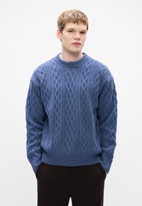 Blauwe kabeltrui met ronde halslijn, met textuurpatronen en ribgebreide manchetten en zoom. Draagt over donkere broeken.
