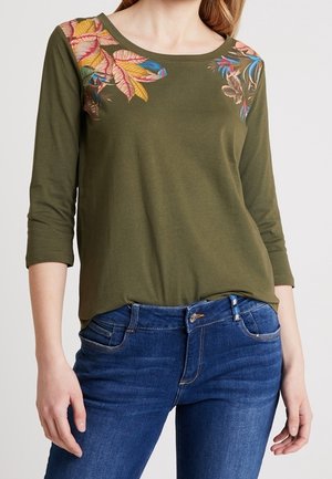 Kvinde iført olivengrøn langærmet top med farverige bladmotiver på skuldrene, kombineret med blå denimjeans.