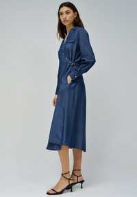 Vestido midi de mezclilla con mangas largas, bolsillos laterales, botones frontales y una cintura ceñida con cinturón. Presenta un dobladillo ligeramente acampanado y una textura suave.
