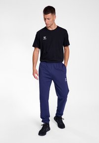 Zwarte t-shirt met logo, gecombineerd met marineblauwe sportbroek met een elastische tailleband. Model draagt zwarte sneakers. Witte achtergrond.