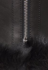 Černý kožený oděv s výrazným stříbrným zipem na jedné straně, který má měkký černý kožich na spodním okraji.