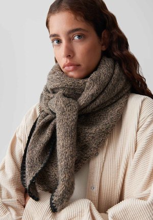 Young woman with long brown hair wearing a thick brown knit scarf and light-colored ribbed jacket, looking directly forward.