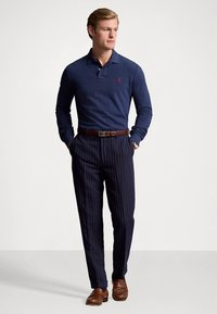 Tamsiai mėlynas polo marškinėliai ilgomis rankovėmis, derinami su dryžuotomis tamsiai mėlynomis kelnėmis ir rudos odos batais. Rudos spalvos diržas ir laikrodis užbaigia aprangą.