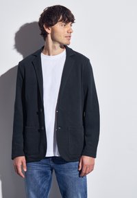 Svart kottblandad kavaj med en strukturerad yta, sjökrage, två framfickor och knappstängning, bärs över en vit t-shirt.