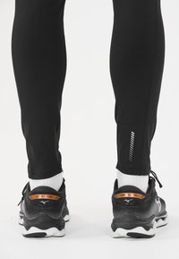 Schwarze, sportliche Leggings mit einer schlanken Passform und reflektierenden Details. Kombiniert mit schwarzen und orangen Laufschuhen und weißen Socken.