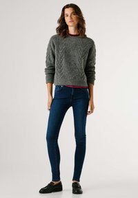 Suéter gris de punto con tejido texturizado, combinado con jeans ajustados azul oscuro y zapatos negros de cordones. Diseño de atuendo simple y casual.