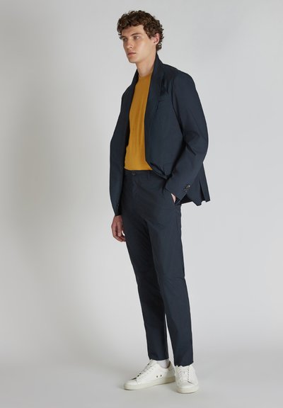 Completo blu navy con blazer su misura e pantaloni coordinati, con una camicia gialla sotto e sneakers bianche, tessuto liscio, vestibilità moderna.