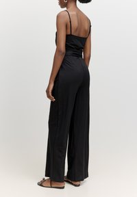Schwarzer Jumpsuit mit dünnen Trägern, tailliertem Oberteil und weiten Beinen. Hergestellt aus glattem Stoff, mit cinchbarer Taille und Seitennähten.