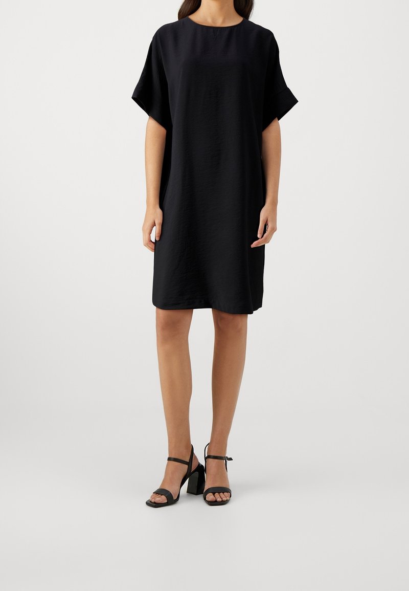 Femme portant une robe noire ample longueur genoux avec manches courtes et des sandales noires à talons hauts, debout devant un fond uni.
