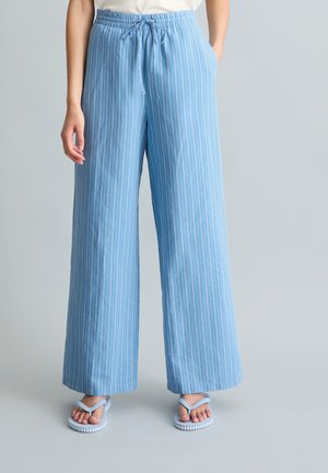 Person trägt locker sitzende hellblaue Hose mit weißen vertikalen Streifen, ein weißes Oberteil und hellblaue Flip-Flops, steht vor einem schlichten Hintergrund.