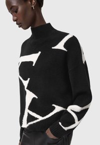 Czarny sweter wełniany z wysokim kołnierzem, zdobiony wyrazistymi białymi abstrakcyjnymi wzorami oraz prążkowanymi mankietami. Miękka struktura z luźnym krojem.