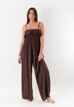 Donna in tuta marrone ampia senza maniche con corpetto arricciato e gambe larghe su sfondo bianco.
