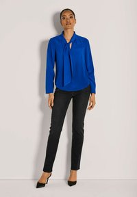 Blaue Bluse mit langen Ärmeln und gebundenem Kragen, kombiniert mit schwarzen, taillierten Jeans. Die Bluse ist glatt und die Jeans haben einen leichten Glanz.