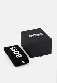 Caja negra con el logo blanco "BOSS" en la parte superior, junto a un bolso de tela negra con el mismo logo. El bolso presenta una franja de contraste en el lado.